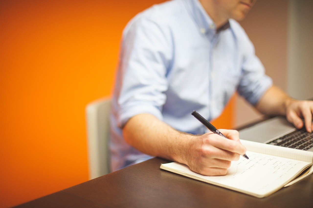A man writing notes in a notebook beside an open laptop